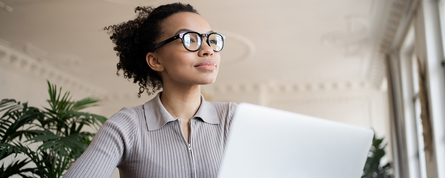 A woman with curly black hair is sitting at her laptop, looking up as if in thought.