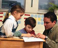 Two children sit at a desk with a teacher, participating in a reading intervention program in a classroom environment.