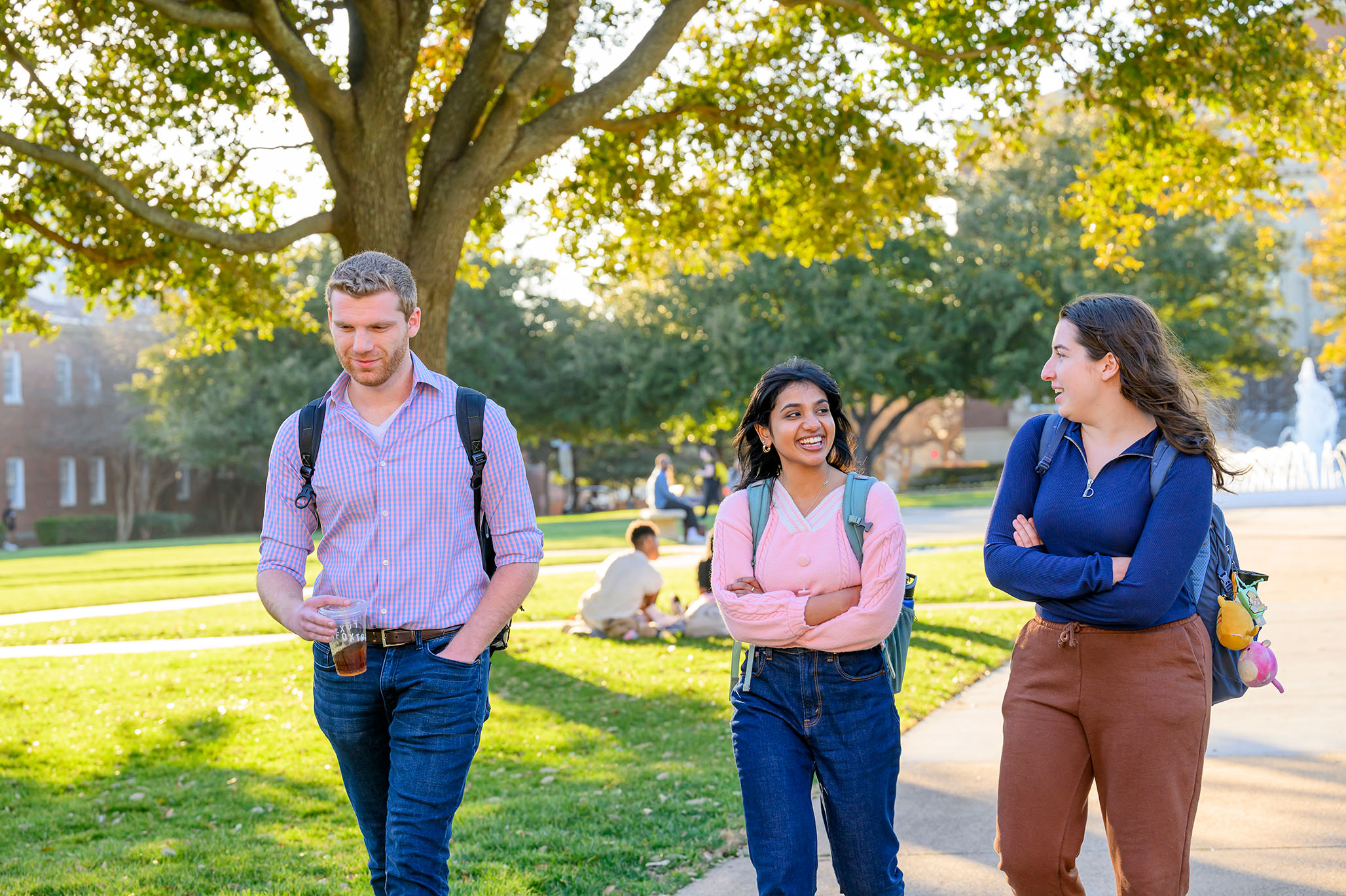 three students walking outside with tree and fountain in background