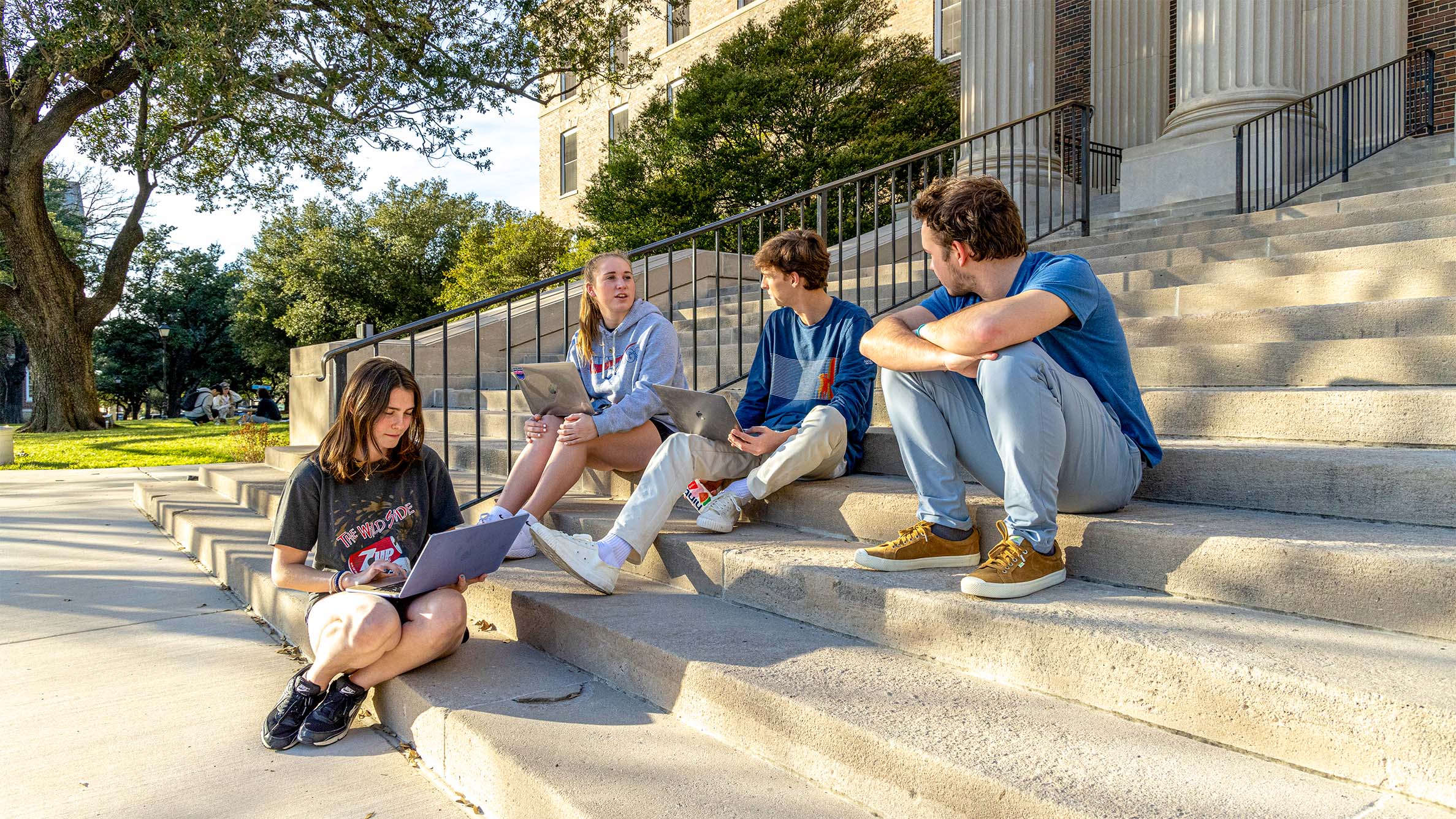 four students sitting on stairs outside of building with laptops talking