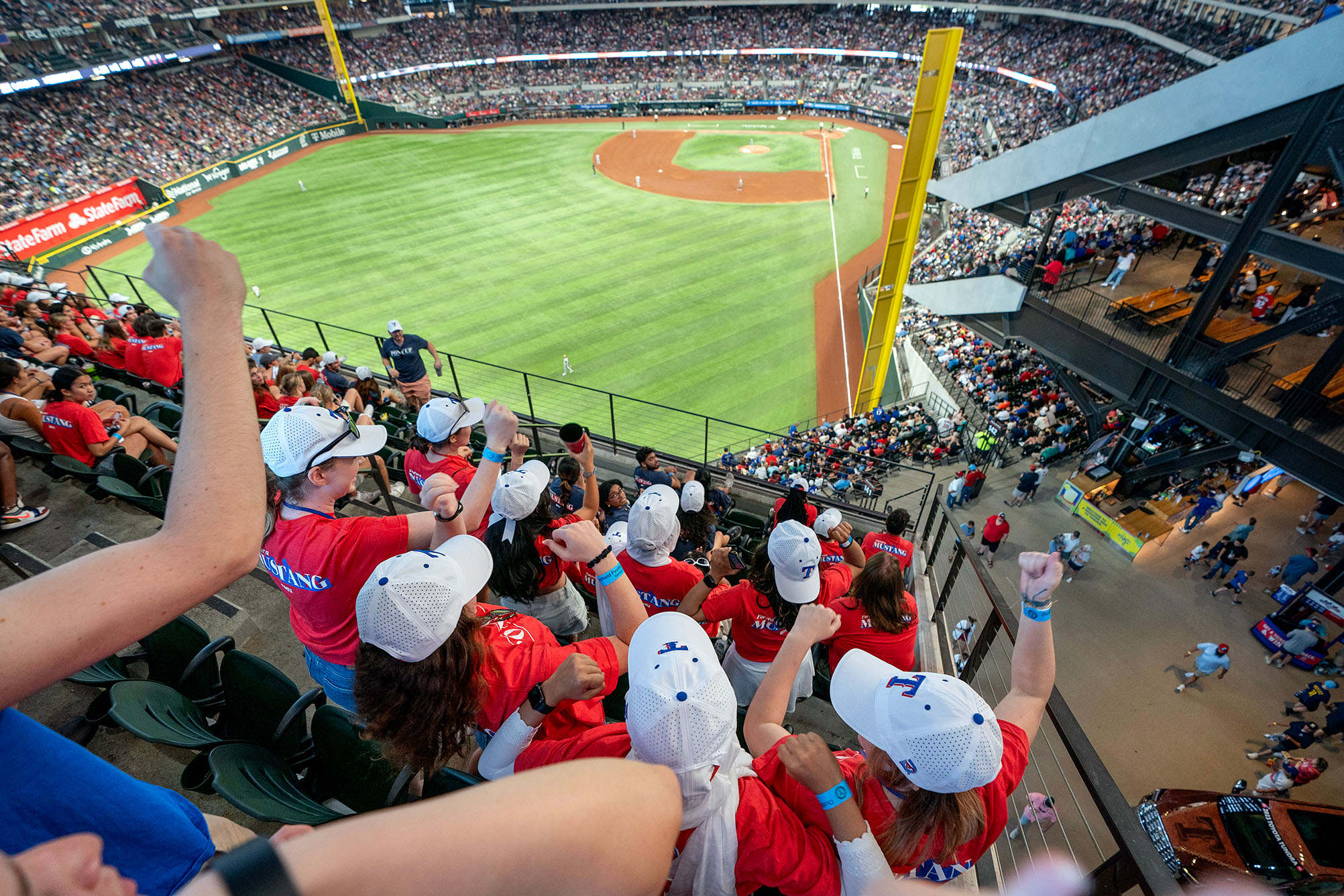 students in red t shirts and white ball caps cheering at Texas Rangers baseball game with field in the background
