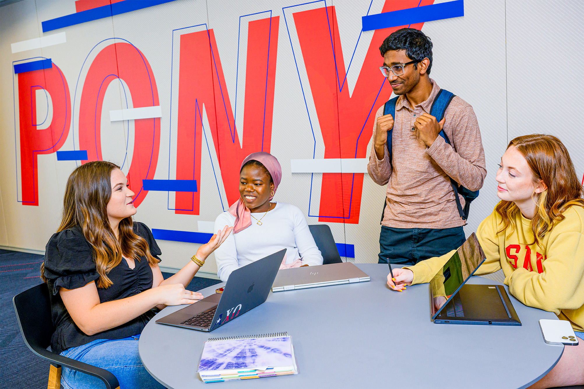 four students sitting at table talking in front of wall, red letters spelling pony on wall